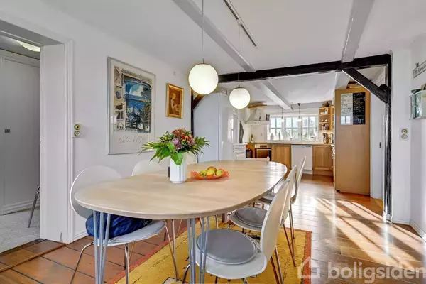 Et langt spisebord står i en lys spisestue. En vase med blomster og en skål med frugt pynter på bordet. Køkkenet ses i baggrunden.