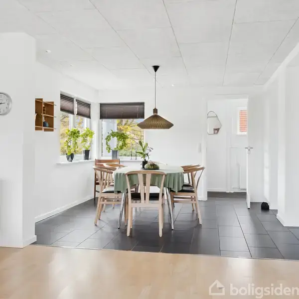 Spisebord med fem stole står på mørkt gulv under en stor hængelampe. Vinduet bagved har planter på karmen. Vægge og loft er lyse. Uret viser klokken halv tre.