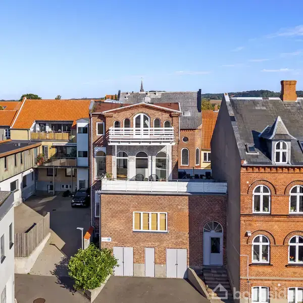 Flere murstensbygninger står tæt ved siden af hinanden i et byområde med blå himmel og få skyer i baggrunden. Etager har forskellige farver og stilarter, omgivet af små gårde og veje.