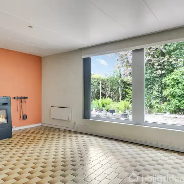 Brændeovn opvarmer et rum med orange væg og ternet gulv. Store vinduer afslører en have med tæt bevoksning udenfor.