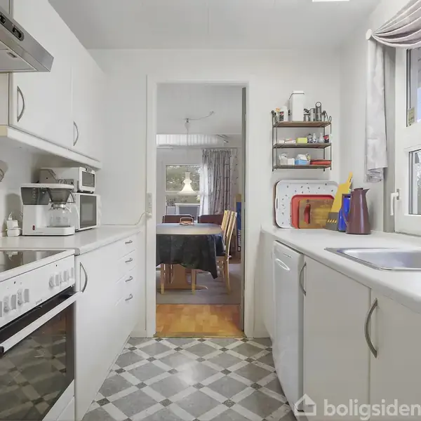 Køkken med hvidt interiør, komfur og vask på hver sin side, handling ingen, med udsigt til en spisestue gennem en døråbning. Mønstret gulv og stort vindue til højre.