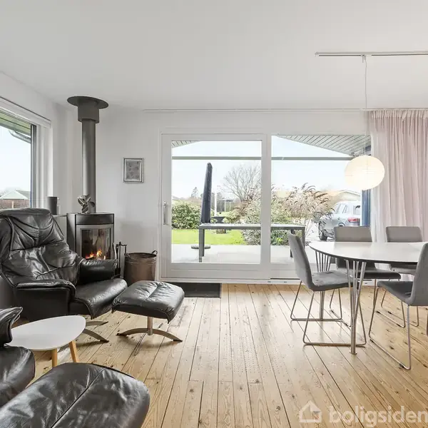 Stue med sorte lænestole og bord, brændeovn med ild, spisebord med stole, store vinduer mod terrasse og have. Trægulve og lyse farver dominerer rummet.