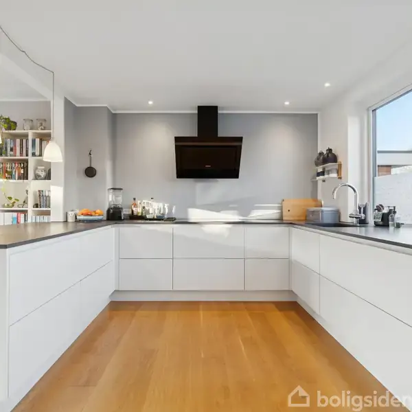 Et moderne køkken med U-formet hvid bordplade, flere skabe og skuffer. Et sort emfang hænger over kogepladen. Køkkenet har trægulv og en hylde med bøger til venstre.