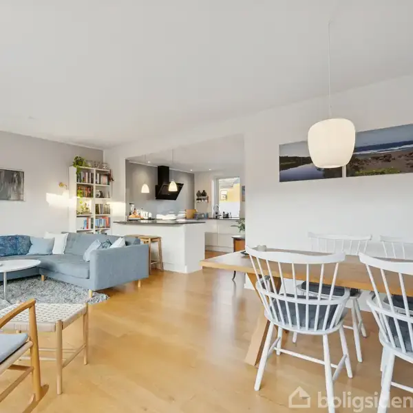 En stue med blå sofa arrangeret rundt om et sofabord, lænestole ved siden af, og et spisebord med hvide stole. Køkkenet er i baggrunden. Indretningen er lys og moderne.