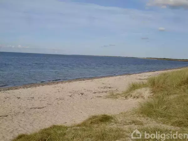 Strand med klart vand og sand, hvor græs vokser ved kanten. Rolig himmel med få skyer skaber en fredfyldt atmosfære.