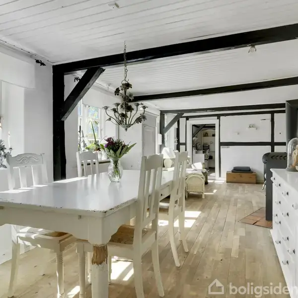 Spisebord med blomster står i lys stue med træbjælker. Der er hvide vægge, trægulv og et stort vindue, der slipper sollys ind. Skab og dekorative genstande står langs væggene.
