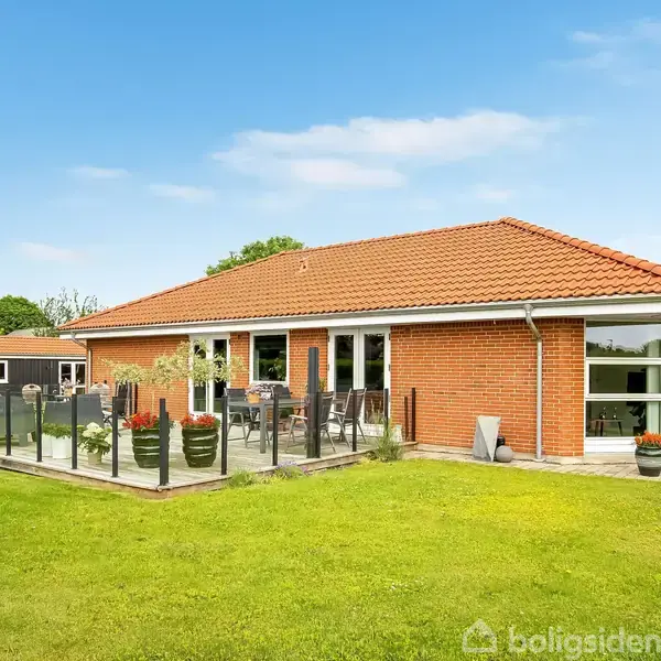Et rødt murstenshus med rødt tegltag står på en velplejet græsplæne. En terrasse med glasrækværk og havemøbler er foran huset, omgivet af grønne planter.