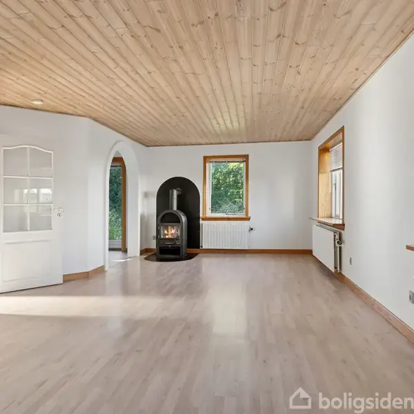 En brændeovn står midt i en lys stue med træloft, lyse trægulve og hvide vægge, omgivet af flere store vinduer og en buet døråbning mod en anden del af huset.