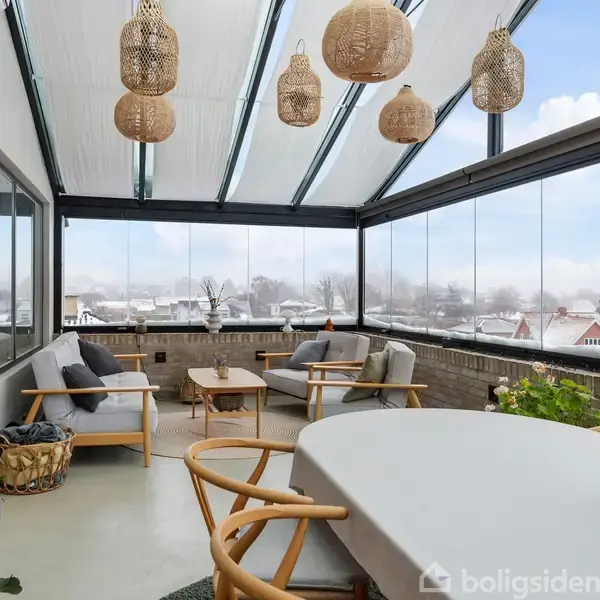 Overdækket terrasse med sofaer og bord i centrum. Der hænger rattanlamper fra loftet. Store vinduer giver udsigt til en sneklædt by med huse og træer i baggrunden.