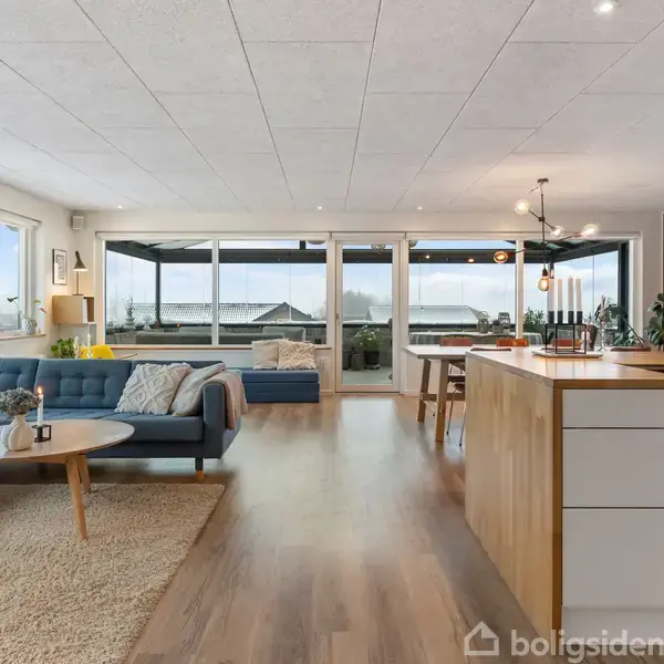 En garderobe med tøj på åbne hylder og en grå skammel. Gennem en glasdør ses et soveværelse med seng og vægmonteret TV. Tregulv og minimalistisk indretning.