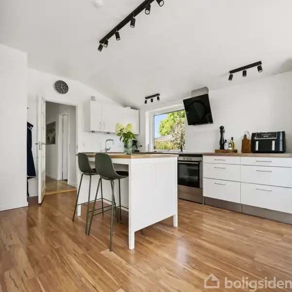 Køkken med hvide skabe og trælaminatgulv. Et lille bord med to stole i midten. Vindue bagved, mikroovn og ovn integreret. Grønne planter pynter på bordet.