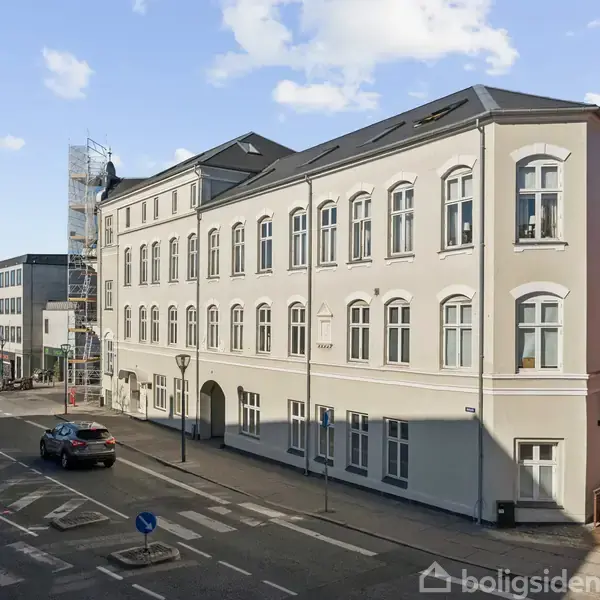 Bygning står statisk ved vejkryds, omgivet af asfaltveje og parkeret bil. Facaden er cremefarvet med mange vinduer, under en blå himmel med få skyer.