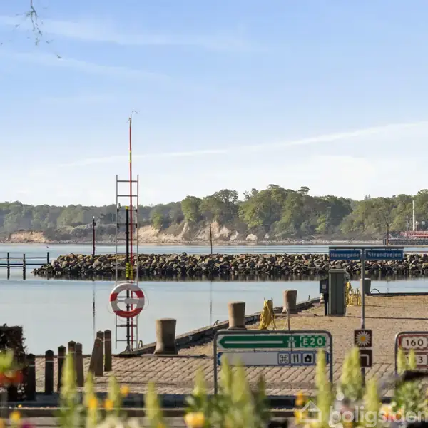 En stille vandhavn med en bølgebryder, omgivet af grønt land. Vejskilte viser mod Odense og Svendborg. Trafiklys og gadelamper er tilstede i forgrunden.