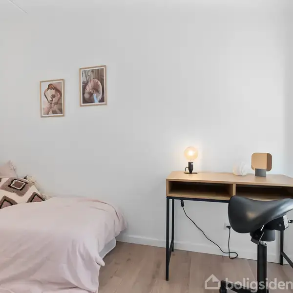 En seng med hvide og brune puder står ved en væg med to billeder. Et skrivebord med lampe og stol står ved siden af sengen i et lyst rum.