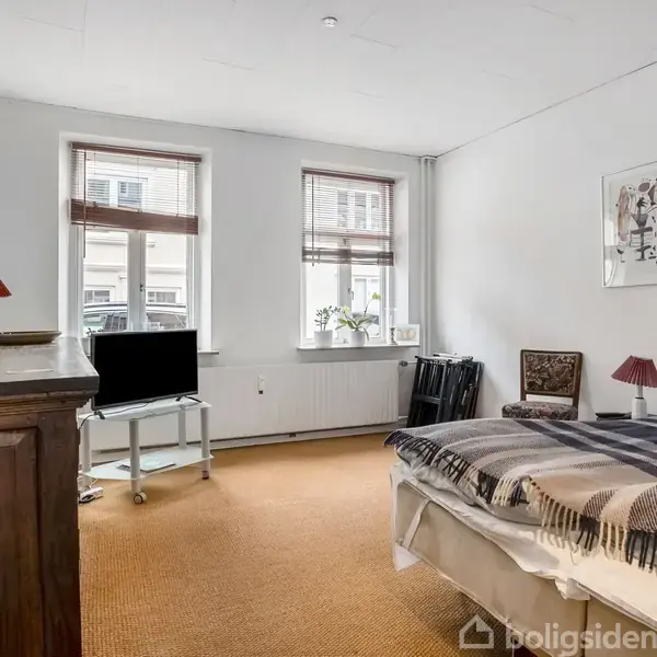 Et soveværelse med en seng dækket af et gråt tæppe til højre, en brun kommode til venstre, en fjernsyn på en lille stander og to vinduer med rullegardiner i baggrunden.