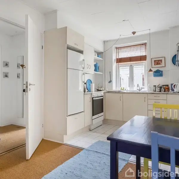 Køkken med hvidt inventar og træbord, hvor forskellige stole står omkring. Et vindue giver lys, og der er køkkenapparater på bordet. Dør åbnes til en anden rummelig gang.