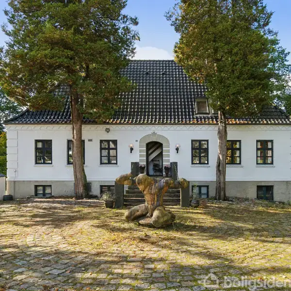 Et hvidt hus med sort tag står blandt høje træer. Foran huset er en brostensbelagt gårdsplads med en stenstatue i centrum.