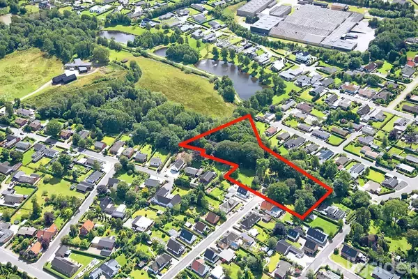 Bebyggelse omkranset af træer set fra oven, hvor et område er markeret med en rød grænse. I baggrunden ses en sø og store bygninger.