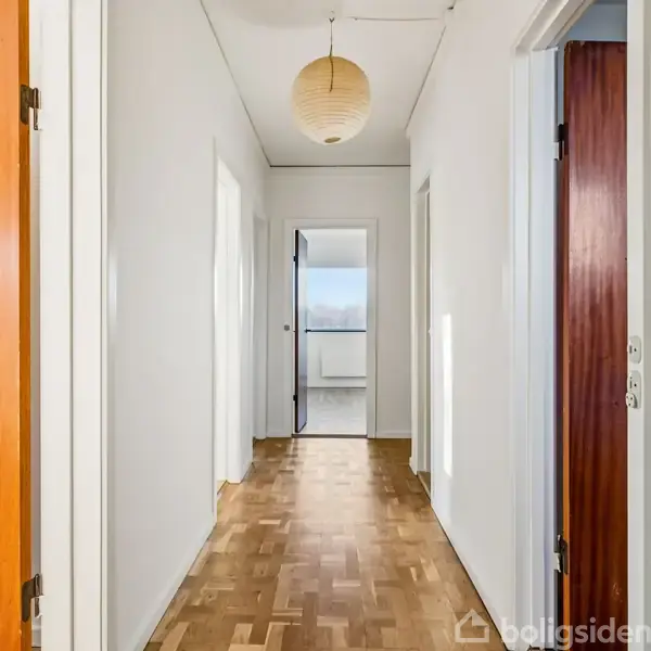 En lang gang med trægulv fører til en dør i enden. Papirlampe hænger fra loftet. Væggene er lyse, og der er flere døre langs gangen.