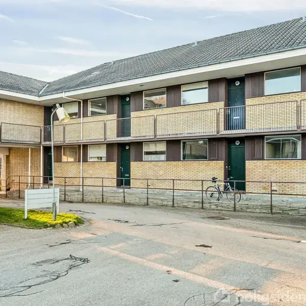 En boligblok med altaner og døre langs anden etage. To biler og en cykel står parkeret foran bygningen, som er omgivet af en asfaltvej og lidt grønne områder.