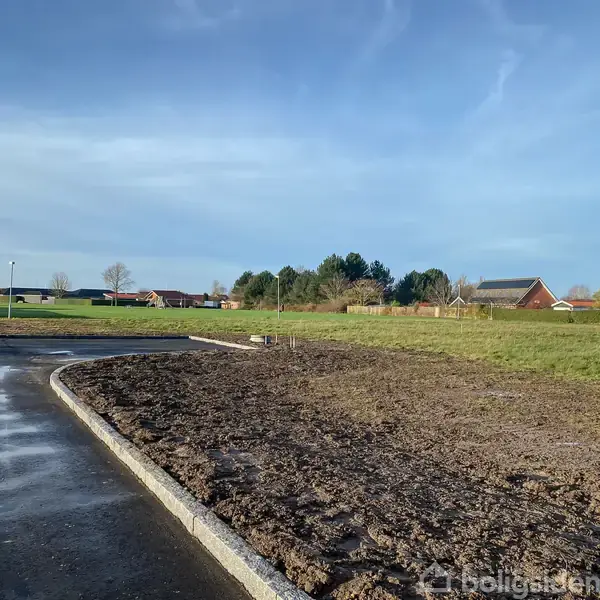 En asfaltvej svinger blødt til venstre nær en bar jordoverflade. I baggrunden ses grønne marker og enkelte huse under en klar blå himmel i et landligt område.