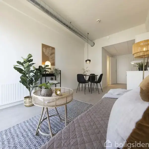 Stue med sofa dækket af quiltet tæppe, lille rundt bord med planter, spisebord med fire stole bagved, vægkunst og moderne belysning. Lyse vægge og minimalistisk indretning.