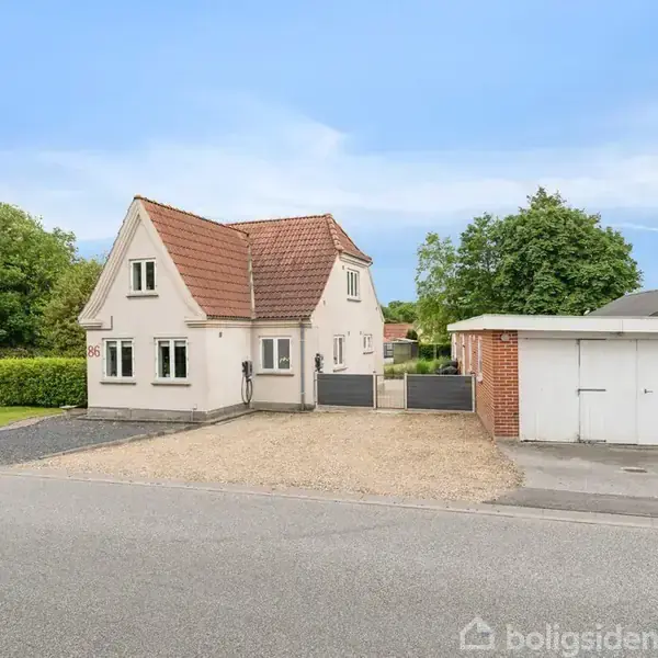 Hvidt hus med rødt tegltag står stille, omgivet af grønne træer og garagebygning. Grusvej foran fører til asfalteret vej.