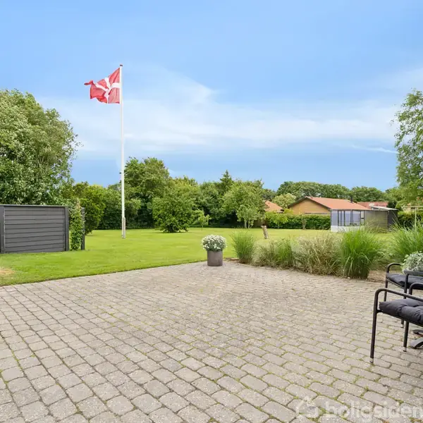 Stenbelagt terrasse med havestole står stille, omgivet af græsplæne og grønne buske. I baggrunden ses et flagstang med Dannebrog, omgivet af træer og en garage.