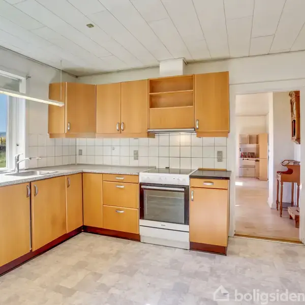 Køkken med lyse træskabe, indbygget ovn og komfur, aktion i et solbelyst rum. Context: Rummet fører til en anden del af huset gennem en døråbning.