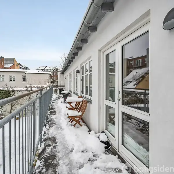 Snedækket altan med et bord og to stole står op ad en hvid bygning med vinduer. Sne dækker jorden, og omkringliggende bygninger ses i baggrunden.