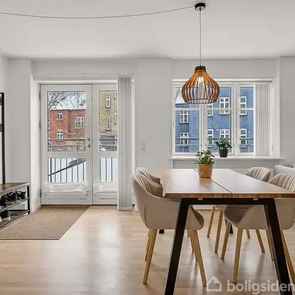 Et spisebord med seks stole står under en hængende lampe i en lys stue. I baggrunden ses en tøjstativ til venstre og sofa til højre med vinduer ud mod bygninger.