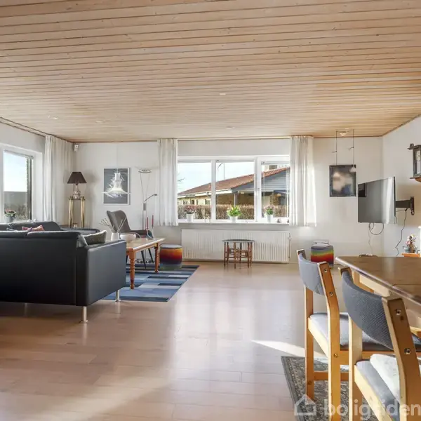 Stue med trægulv og loft, indeholder sorte sofaer, stort træbord med stole, fladskærms-tv på væggen og store vinduer med hvidt gardin, der lader lys strømme ind.