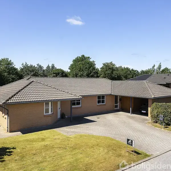 En enkeltstående, lysebrun murstenshus med mørkt tegltag. Huset har en integreret carport og ligger i et grønt boligområde. Nummeret "42" ses ved indkørslen. Græsplæne foran huset.