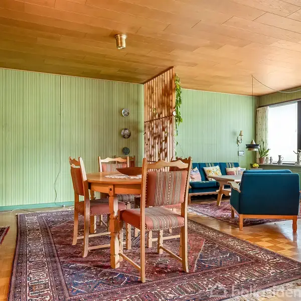 Et spisestueområde med et bord og stole står på et tæppe. I baggrunden ses en stue med blå sofaer nær vinduet. Rummet har grønne vægge og trægulv.
