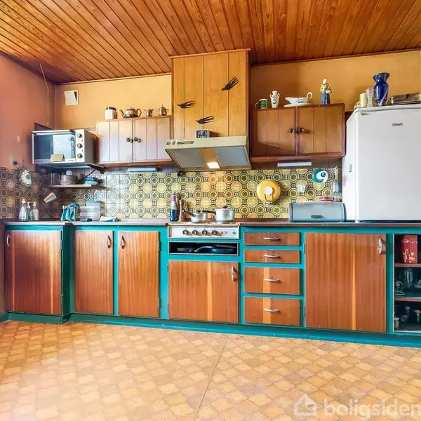 Køkken med træskabslåger, retro flisemønster og blågrønne detaljer. Der står en mikrobølgeovn ovenpå skabene, et køleskab ved siden af, og en ovn i midten. Hyggelig atmosfære.