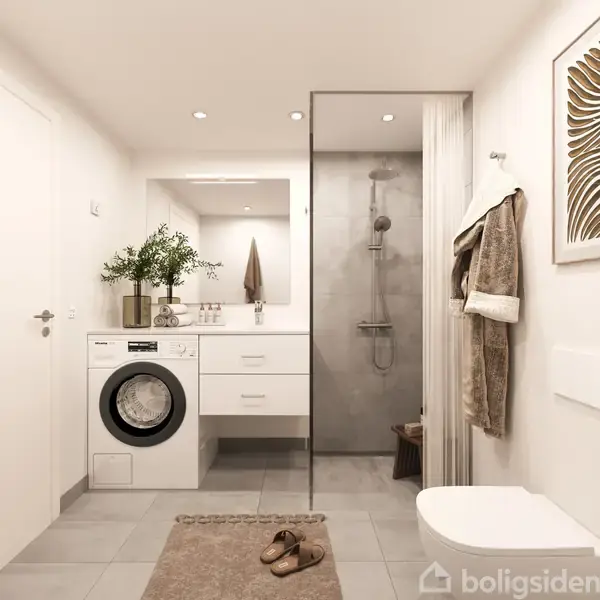 Vaskemaskine under hvid vask med spejl, planter og rullet håndklæder ovenpå. Bruseniche med glasvæg til højre. Beige håndklæde hænger på en krog ved siden af.