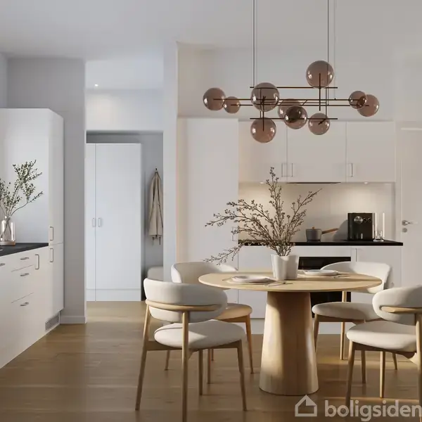 Et rundt spisebord med en vase fyldt med grene står midt i et lyst, moderne køkken. Omkring bordet er fire polstrede stole. Køkkenet har hvide skabe og en stor vindueslysning.
