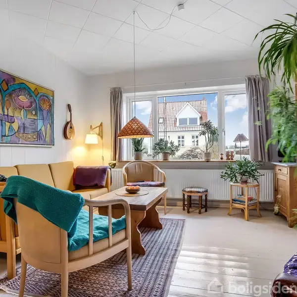 Stue med træmøbler og farverig kunst på væggen. Lampe hænger over sofagruppe. Store vindue med planter og udsigt til et hus. Grønne planter skaber et hyggeligt miljø.