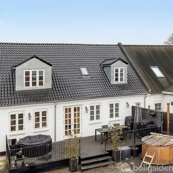 Hvidt hus med sort tegltag, to døre, og flere vinduer. Dobbelt repos med grill og udendørs badekar. Placeret i en have med murstensbelægning.