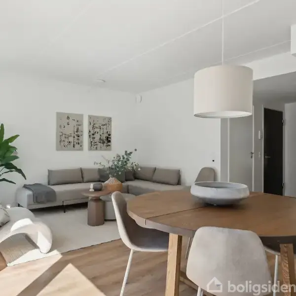 En lys dagligstue har et rundt spisebord med fire stole og en sofa i baggrunden. Rummet er minimalistisk indrettet med grønne planter, kunstværker på væggen og træmøbler.