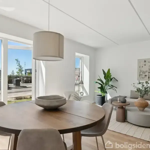Rundt spisebord med beige stole står i en moderne, lys stue. En sofagruppe, grønne planter og kunst på væggen udgør indretningen. Store vinduer viser en bygning udenfor.