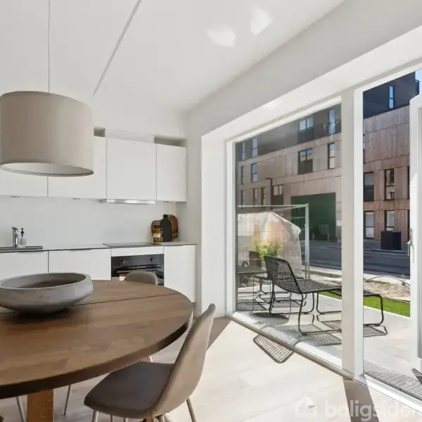 Et rundt træbord med stole står i et lyst køkken. Store vinduer viser en moderne bygning udenfor. En loftlampe hænger over bordet. Ingen tekst er til stede.