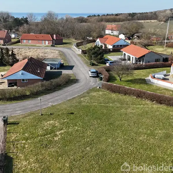 Huse med røde tage ligger langs en snoet vej i et grønt landskab, med havudsigt i baggrunden. Det er en stille beboelsesområde med spredte træer og buske.