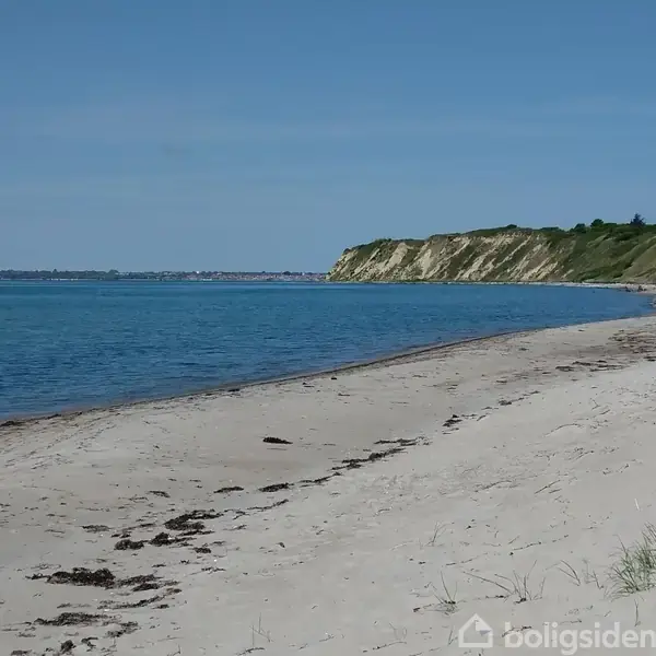 En sandstrand strækker sig langs kysten med klart blåt hav ved siden af. Grønne klipper former baggrunden under en skyfri himmel.