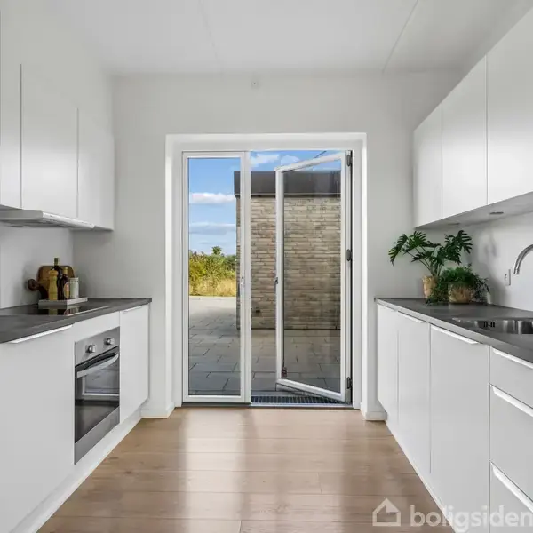 Toilet og brusebad i et lyst badeværelse. Stor spejl over hvid vask med skab, grøn plante og sorte beholdere. Beige gulvfliser og blå håndklæde på væggen.
