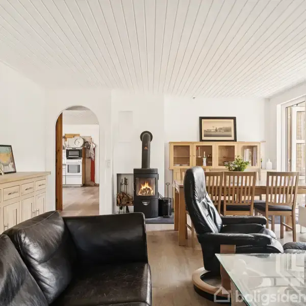 Sort lædersofa og lænestol står omkring et glasbord i en stue med brændeovn. Et spisebord med stole er ved siden af store vinduer, der giver udsigt til haven.
