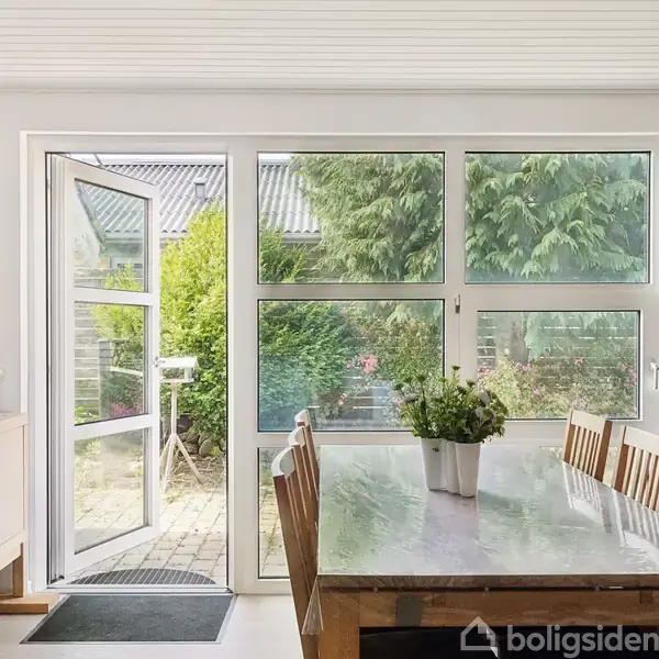 Et spisebord med stolene står ved lange vinduer, der kigger ud til en grøn have. En dør står åben mod terrassen, og en plante pynter bordet.