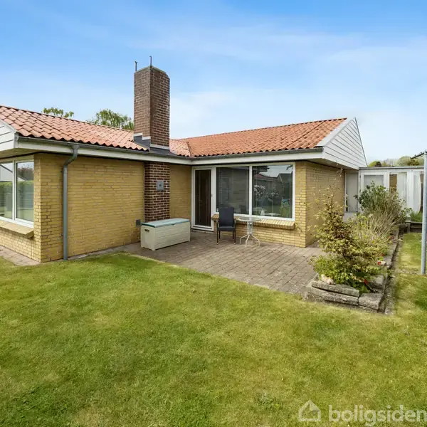 Et gult murstenshus med rødt tegltag står stille i en velplejet have med fliseterrasse. Store vinduer og en skorsten ses, omgivet af træer og en græsplæne under en klar blå himmel.
