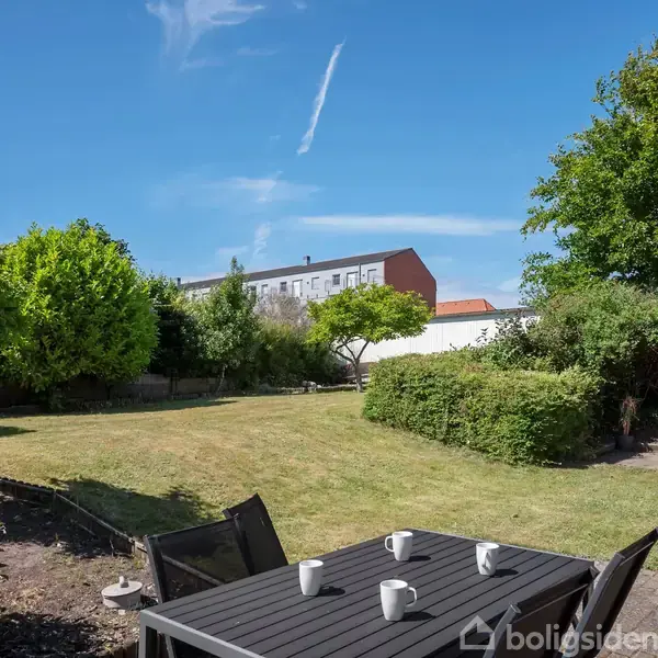 Et sort havebord med fire hvide krus står på en terrasse. Bagved strækker en græsplæne sig ud, omgivet af forskellige grønne buske og træer under en klar blå himmel.