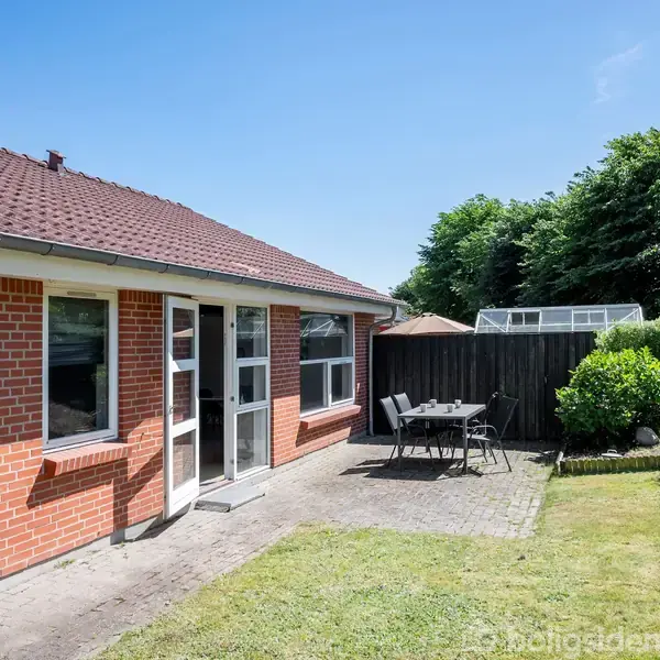 Rød murstenshus med rødt tegltag, åbne vinduer og dør, står i en have med græsplæne, flisebelagt terrasse, sort havebord og stole. Omgivet af buske og træer under en blå himmel.